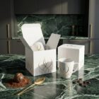 Espressotasse „Kaffeeduft“, elegante Designer-Espressotasse aus Porzellan mit feiner, goldfarbener Illustration von NONOS, Espressotasse mit Untertasse, Kaffee und Praline auf grünem Marmortisch, stilvolle Detailaufnahme.